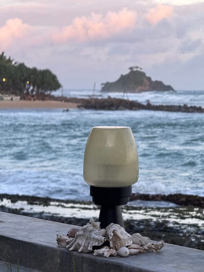 A lamp with a ribbed, translucent globe is mounted on a dark pole. Attached to the pole near the globe are several seashells and pieces of coral, some appearing weathered. The lamp and shells are positioned on the right side of the frame, resting on a gray concrete ledge.
Beyond the ledge, a dynamic ocean scene unfolds. The water is a turbulent mix of blues and whites, indicative of crashing waves. A rocky breakwater runs parallel to the shore, separating the rough sea from a calmer, sandy beach visible in the background. Trees line the edge of the beach, with some lights twinkling amongst them, suggesting a settlement or resort. In the distance, a small, tree-covered island rises from the sea.
The sky above is filled with soft, muted clouds, tinged with the pale colors of either dawn or dusk. The overall atmosphere suggests a serene, yet wild, coastal environment. The image appears to be taken outdoors, likely in the late afternoon or early morning, given the soft light and cloud colors. The location is identified as Ahangama, Sri Lanka. No people are visible in the image, and there is no visible text.