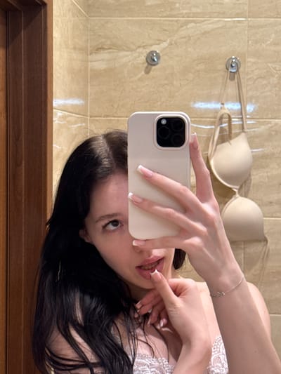 This is a selfie taken by a young woman with long, dark hair. She is holding a smartphone in her right hand, angled to capture her reflection in a mirror. The background appears to be a bathroom with beige tiled walls. A wooden door frame is visible on the left. Two beige bras are hanging on hooks behind her. Her left hand is near her mouth, and she is wearing a light-colored lacy top and a delicate silver bracelet on her left wrist. The image does not provide any specific information about the city of Khimki, Russia.