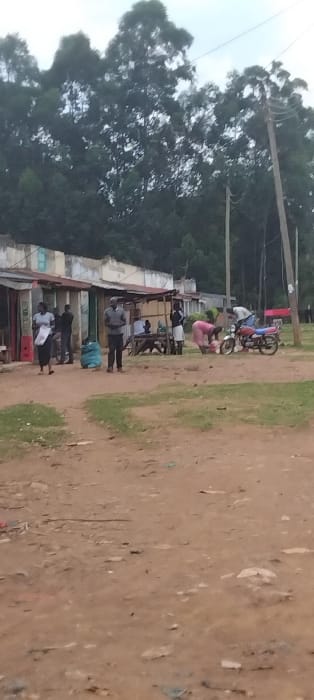 The image shows a dirt road in front of several buildings, presumably shops, in the town of Lwandeti, Kenya, located at latitude: 0.563, longitude: 34.807. There are several people present, including three men standing by the shops, two women in pink clothing standing on the road, a man and a woman standing near a motorcycle, and a woman in a blue dress sitting on the ground. There is a motorcycle parked on the side of the road, and a few other objects such as bags and chairs can be seen scattered about. The image is captured from a high angle, looking down at the scene. The buildings are made of concrete and have a corrugated metal roof. The scene is set against a backdrop of dense, green trees.