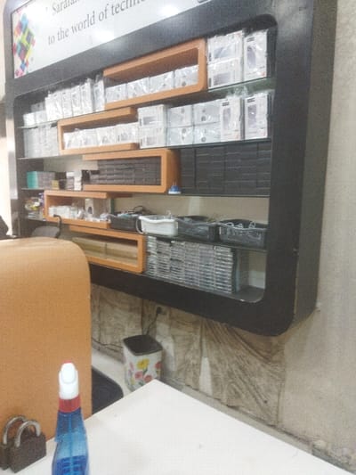 The image depicts the interior of what appears to be an electronics or mobile phone shop in Nuseirat, Palestinian Territory. The central focus is a large, dark-colored wall-mounted shelving unit with integrated light brown/orange rectangular compartments, displaying various electronic goods and accessories.

The top three shelves are primarily filled with stacks of white boxes, many individually wrapped in clear plastic, consistent with packaging for mobile phones, chargers, or other small electronic devices. Some of the rectangular compartments on these shelves hold dark, uniform items. The bottom shelf contains black plastic baskets filled with miscellaneous smaller items, possibly components or accessories. To the far left of the bottom shelf, stacks of silver-colored cases or discs are arranged horizontally. A small blue and white electrical adapter or device is visible on the third shelf from the top.

A sign mounted above the shelving unit features partially visible text "Sarala..." and "to the world of tech...", along with a colorful square pattern. The wall below the shelving unit exhibits a textured, somewhat distressed appearance, possibly due to water damage or a decorative finish.

In the immediate foreground, a white table surface is visible. On this table, a blue plastic spray bottle with a white nozzle and a red cap is positioned. To the left of the spray bottle, a dark-colored padlock with two silver shackles is present. Partially visible in the lower left, a large, tan-colored upholstered object suggests the back of a chair or seating. A white waste bin with a floral pattern (yellow, red, green flowers) is situated on the floor beneath the shelving unit. No individuals are visible in the scene.