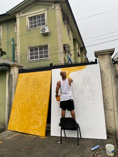 A man stands on a chair, painting a large canvas in Ado, Nigeria. The canvas, leaning against a wrought-iron fence, depicts figures sketched in pencil and partly painted in yellow. A second, similar canvas, painted yellow, lies adjacent. The man wears a white tank top and dark shorts. Painting supplies are on the ground nearby. A green, multi-story building with white-framed windows and decorative molding is in the background. Overhead, power lines are visible against a grey sky.