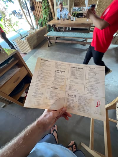 The image depicts a person holding a menu at Lilla Pantai, a restaurant in Denpasar, Indonesia. The menu is open, displaying various food and drink options under categories such as "Breakfast," "Smoothie Bowls," "Sandwich," "Burger," "Small Bites," "Soups," "Salads & Bowls," "Pasta," and "Vegetarian." The background shows a rustic and airy dining area with wooden furniture, wicker seating, and lush greenery, characteristic of many establishments in Denpasar. An older man is seated at a table in the background, while another person in a red shirt is partially visible walking past. The lighting suggests it is daytime, with sunlight filtering into the space.