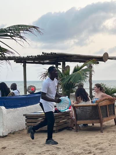 At an outdoor cafe on a sandy beach under a cloudy sky, a man in a white shirt and black pants holds a blue and white circular decoration. Two women sit on a wooden bench behind him. Another person sits near a blue and white decorative wall. Palm fronds and a thatched roof structure are also visible.