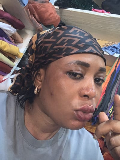 A close-up, eye-level selfie depicts an adult female individual with a dark complexion, positioned in an indoor commercial setting. She is wearing a light grey crew-neck t-shirt and a black and brown patterned headwrap featuring a repeating "F" logo, indicative of the Fendi brand. Her dark, curly hair is visible beneath the headwrap on her left side. Her face exhibits a shiny sheen, possibly from sweat or oil, and her full lips are pursed in a pouting expression. Her eyes are lined with dark makeup and gaze directly at the camera. A gold-toned hoop earring is visible in her left ear. Her right hand is raised, with the index finger slightly curled upward.
The background suggests a fabric or textile shop, likely located in Sabon Tasha, Nigeria. To the left, stacks of various rolled or folded fabrics are visible, including yellow, light green, maroon, and white textiles, some on shelves. A white fabric with a red "F" logo (Fendi) is also present. To the right, multiple vertical strips of multi-colored fabric, possibly ribbons or samples, hang. The upper background shows additional shelves containing crumpled and folded fabrics of different colors, including an orange-brown and a green sequined material. The environment appears functional and densely packed with merchandise.