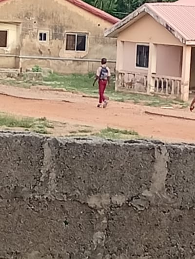 A partially obscured view shows a dirt road in Adankolo, Nigeria. A person wearing red pants and carrying a backpack walks along the road. A concrete wall occupies the foreground. In the background are two buildings, one faded yellow and one a newer peach color. The sky is not visible.
