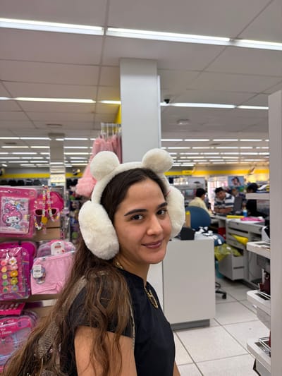 A woman wears white, fluffy bear-ear earmuffs indoors. She smiles slightly, positioned next to a shelf displaying pink merchandise, including "Hello Kitty" items. Bright fluorescent lighting illuminates the scene. In the background, blurred figures suggest a store environment, further enhancing the retail context.