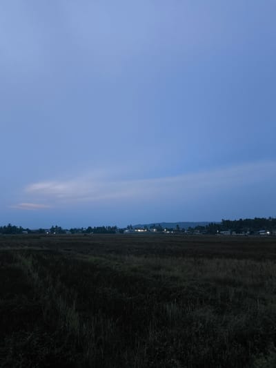 The image captures a rural twilight scene in Bireuenmeunasahblang, Indonesia. The sky is a deep, gradient blue, transitioning from a lighter hue near the horizon to a darker shade overhead. Faint, wispy clouds are visible on the horizon, tinged with a soft pinkish-orange light from the setting sun. Below the sky, a strip of landscape stretches across the frame. This strip is dominated by a dark, fallow field in the foreground, its texture a mix of dry stubble and shadowy vegetation. In the mid-ground, a line of trees and buildings forms a village or settlement, indicated by scattered lights from houses and streetlamps. The distant background features rolling hills, partially obscured by the twilight. The overall impression is one of quiet dusk in a rural Indonesian setting.