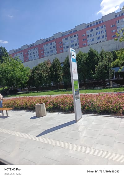 This is a medium shot photograph taken on a sunny day, likely in the late morning or early afternoon, given the strong shadows. The scene appears to be a public outdoor space, possibly a bus stop or transit area, in Warsaw, Poland.

In the foreground, a paved walkway made of light gray square tiles is visible, with a textured tactile paving strip running across it. To the left, a light brown wooden bench is partially visible, with a person in a light brown jacket and blue jeans sitting on it. A concrete trash receptacle, with an octagonal shape and weathered texture, stands to the right of the bench, casting a distinct shadow on the pavement.

Behind the walkway and bench, a line of vibrant pink flowering bushes creates a colorful border. Further back, a grassy area is dotted with various trees, some with lush green foliage and others showing signs of seasonal change with brown leaves. In the mid-ground, a modern bus stop sign with a blue and white bus icon stands prominently. Text on the sign is partially visible, including "Warta Tanie Pojeżdzić," which suggests information about transportation or travel offers.

Dominating the background is a large, multi-story residential building with a distinctive architectural style, featuring a pink band across its upper floors and numerous windows. The sky is bright blue with a few scattered white clouds, indicating clear weather.

The bottom of the image contains a timestamp and camera information: "NOTE 40 Pro" and "2026.06.17 12:52," along with camera settings "24mm f/1.75 1/207s ISO50." The overall impression is one of a typical urban environment on a pleasant day.