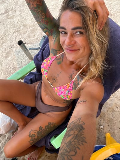 A high-angle, full-body selfie shows a woman with blonde hair, tan skin, and numerous tattoos lying on a beach. She is wearing a floral print bikini top and brown bikini bottoms. Her right arm is bent, with her hand resting on her head. Her left arm is outstretched, showing tattoos on her arm and shoulder. The woman is lying on a beach chair covered with a dark blue towel, with sand visible in the background. A yellow object, possibly part of a beach umbrella, is visible on the right side of the image. The lighting suggests it is daytime, likely sunny. The woman has a slight smile on her face, and her eyes are open and looking towards the camera. Visible tattoos include floral designs, geometric patterns, and script on her arms and chest. There is a nose ring and eyebrow piercing. The setting is a beach, implied by the sand and the presence of beach furniture. The overall mood is relaxed and casual, typical of a beach vacation.