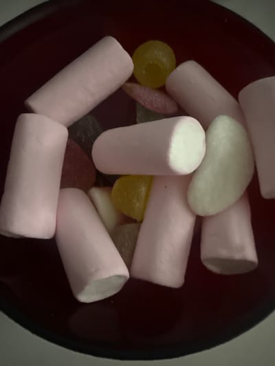 The image shows a close-up, overhead view of a dark red bowl filled with an assortment of candies. The candies include several pink cylindrical marshmallows, some yellow spherical candies, a light green bean-shaped candy, and a few dark red and black gummy candies. The lighting is dim, and the focus is slightly soft, giving the image a candid and slightly hazy appearance. There is no indication of a specific location within the city of Valencia, Spain, as the image focuses solely on the contents of the bowl.