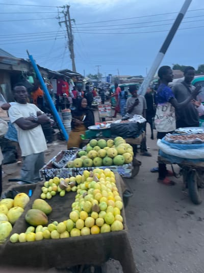 The image depicts an outdoor market scene in Gura Top, Nigeria, under a cloudy sky. In the foreground, a man in a light-colored t-shirt and gray pants stands with his arms crossed, facing left. To his right, a stall displays a large quantity of light-green and yellow fruits, likely guavas, arranged in rows. Behind this stall, another stall is visible with more fruits. Several people are milling about in the background, with market stalls and structures lining the street. A blue pole stands near the center of the frame, and power lines crisscross the sky. The ground appears to be paved. The overall impression is a busy street market environment.