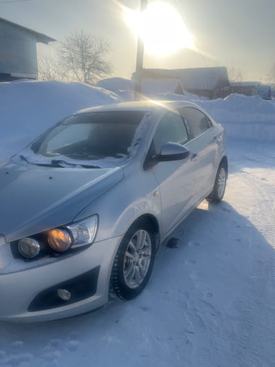 A silver Chevrolet Aveo (T300 generation) sedan is parked on a snow-covered surface under bright, low-angle sunlight in Kozlovka, Russia. The vehicle's front left quarter is prominent, with snow accumulated on its hood, windshield, and wipers. The passenger-side (image-left) front turn signal or parking light is illuminated orange. The car is equipped with multi-spoke alloy wheels and winter tires. Deep snowdrifts line the immediate foreground and background, obscuring parts of surrounding structures, which appear to be residential or utilitarian buildings. A utility pole stands upright, and bare trees are visible in the distant background. Long shadows are cast across the snow, and sunlight creates a strong glare in the upper right portion of the frame, indicating a clear, cold winter day. No individuals or active interactions are visible in the scene.
