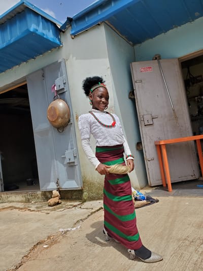 A young woman stands in front of a light blue and tan building, with a metal door visible. She is wearing a white long-sleeved top with intricate lace detailing, a colorful beaded necklace, and a skirt with maroon and green diagonal stripes. She has a blonde wig on, and is smiling at the camera. Beside her, a broom leans against the building. The ground is concrete with some cracks. The image is taken outdoors, with what appears to be a bright, sunny day, casting shadows. A metal pot hangs from the side of the building. In the background, there is a large, gray metal door or panel with a red "WINNER" sticker. The scene appears to be in Gura Top, Nigeria.