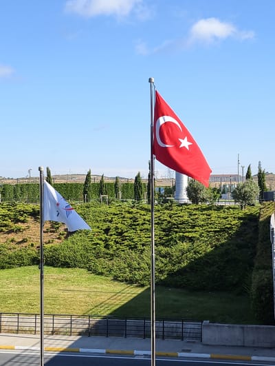 This media file is a photograph taken outdoors on a clear day with a bright blue sky and a few white clouds. The scene features two flags on flagpoles, with a lush green landscape in the background.
The prominent flag on the right is the Turkish flag, a red ensign with a white star and crescent. It is waving in the wind. To the left of the Turkish flag is another flag, predominantly white, with a red star and some blue and white emblems. This flag also appears to be in motion, as if caught by the breeze.
The background consists of a tiered landscape covered in dense green bushes, reminiscent of manicured hedges. In the distance, there are trees, some power lines, and industrial structures, including a tall, white, cylindrical building with a spiral design. The ground in the foreground shows a grassy area and a paved pathway with yellow and white markings. A black metal fence runs along the edge of the landscape.
The bright sunlight suggests it is daytime, likely late morning or afternoon. The shadows cast on the grass indicate the sun's position. The setting appears to be an open area, possibly a park, a campus, or an industrial complex. There is no visible text in the image. The overall impression is one of a clean, modern, and well-maintained environment.