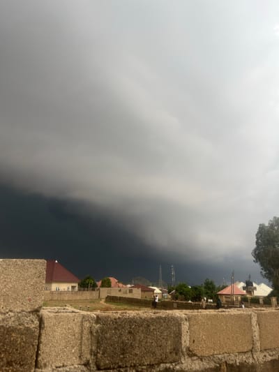 In Barakin Akawo, Nigeria, a dramatic sky looms over a residential area. The foreground is dominated by rough, textured concrete blocks, suggesting a construction site or unfinished building. Beyond this barrier, a line of houses with red and white roofs are visible, nestled amongst sparse vegetation. Several individuals are scattered amongst the houses, some appearing to be walking or standing. In the distance, cell towers rise above the treeline, and a distant hill can be seen. The most striking element is the immense, dark storm cloud that fills the upper two-thirds of the image, casting a heavy, ominous shadow over the landscape. The contrast between the bright sky at the top and the dark, brooding storm cloud is stark, creating a sense of impending weather.