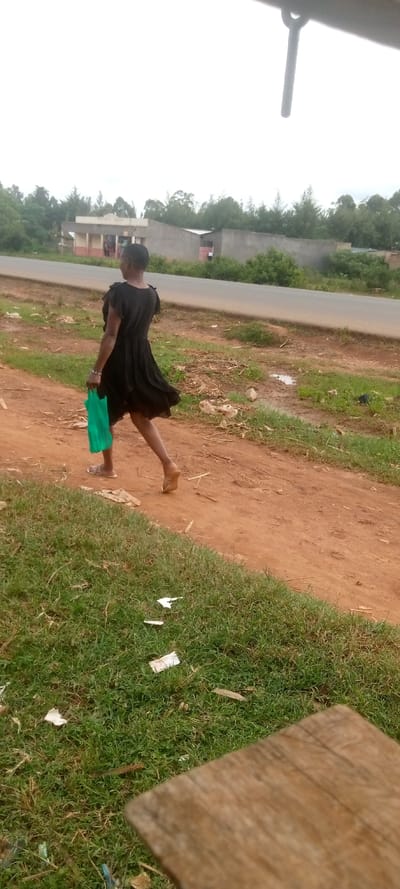 A woman wearing a black dress and sandals walks on a dirt road while carrying a green plastic bag. The road is flanked by green grass and a gray concrete wall, a small structure is visible in the background to the left of the scene. The road has a paved surface. The woman is walking from right to left. The woman is in focus, the background is out of focus. The scene takes place in a rural area of Kivaywa, Kenya. The road is lined with trees on the right side. The scene is overcast and grey. There is a wooden object and debris in the foreground. There are a few small puddles on the road. The scene is taken from a low angle, looking up at the woman. The image quality is poor.
