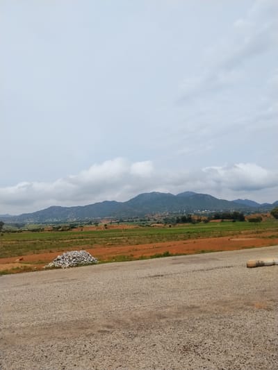 This is a wide-angle, eye-level photograph capturing a rural landscape under a cloudy sky. The foreground is dominated by a gravelly road or construction area, with a pile of grey stones to the left. Beyond the road, a field of reddish-brown soil transitions into green agricultural land, dotted with small white birds. In the mid-ground, a chain of low, green-covered mountains stretches across the horizon. Small clusters of white buildings are visible on the slopes of the mountains, suggesting a rural village. The sky is mostly overcast with scattered white clouds. The image appears to be taken in the vicinity of Dorowa, Nigeria, based on the characteristic landscape of the region, which often features rolling hills, agricultural fields, and scattered settlements. There are no people or specific actions depicted in the scene. The overall impression is one of a tranquil, undeveloped rural environment.