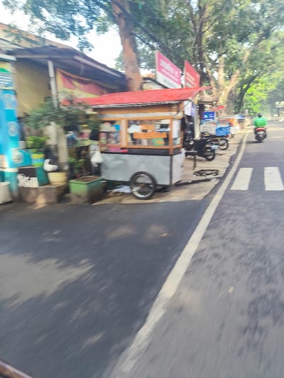 The image shows a street scene in Bandung, Indonesia, taken from a moving vehicle. The camera is focused on a street cart with a red roof and a gray body, located on the right side of the street. The cart is parked and appears to be selling food. The street has a white line down the middle, and there is a pedestrian crosswalk in the distance. There are trees lining the street on both sides. A motorcycle is driving down the street. The image is out of focus and motion blurred. The coordinates provided place the scene in a residential area near a school. The image suggests a busy street in an urban environment.