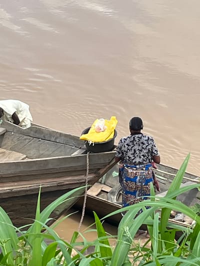 The image portrays a tranquil river scene in Idah, Nigeria, featuring two individuals in traditional wooden boats amidst lush green vegetation. The calm river water, a muted brown or beige, occupies the left side of the frame.
In the foreground, a person is partially visible in a wooden boat, seen from behind, wearing a light-colored or white garment. Further back, another individual, also facing away from the camera, is situated in a second wooden boat. This person wears a black and white patterned short-sleeved top and a vibrant blue fabric wrap or skirt adorned with gold and brown patterns, typical of traditional attire. Their head is covered by a dark cap, and they are in a bending or squatting posture, seemingly engaged in an activity within the boat.
A thick rope connects the two boats, suggesting they are moored together. A bright yellow object, possibly a bag or piece of clothing, rests near the bow of the front boat. The right side of the scene is framed by abundant, tall green foliage, likely reeds or grasses, characteristic of a riverside environment. The natural daylight suggests it is daytime. No text is visible in the image. The scene captures a quiet moment of daily life on the river.