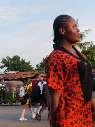 A medium shot captures a woman in an orange and black floral print top standing on a city street. Her hair is styled in braids pulled back from her face, and she looks to her right. In the background, three students in school uniforms walk across the street. A green and brown three-wheeled vehicle is parked nearby. Trees and building roofs appear in the background under a partly cloudy sky. The location is consistent with a busy street in Aba, Nigeria.