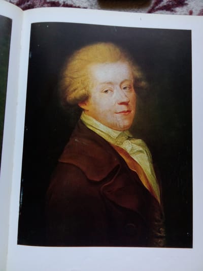 The image is a painting of a man with fair skin, blond hair styled in a powdered wig, and a light blue gaze. He is wearing a dark brown coat over a light yellow shirt with a ruffled collar and a sash. The painting is set against a dark background. The painting is presented within a book.