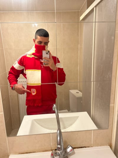 A person in a red Adidas tracksuit and a red and white scarf with a logo is reflected in a bathroom mirror. The person holds a phone, presumably taking the picture. A white sink with a silver faucet is to the right. Beige tiles cover the wall. The mirror shows a toilet and a partial view of the bathroom. The person points at the scarf's logo.