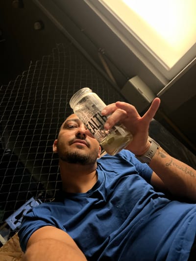 In an indoor setting, a man with short hair and a beard, wearing a blue t-shirt, is visible from the chest up, looking upwards. He is holding a clear glass jar in his right hand, with his thumb extended. The jar contains a green substance. The background includes a black chain-link fence and a bright overhead light fixture. The man has tattoos on his right arm. The image is a low-angle shot. There is no indication of the city of Sevlievo, Bulgaria.