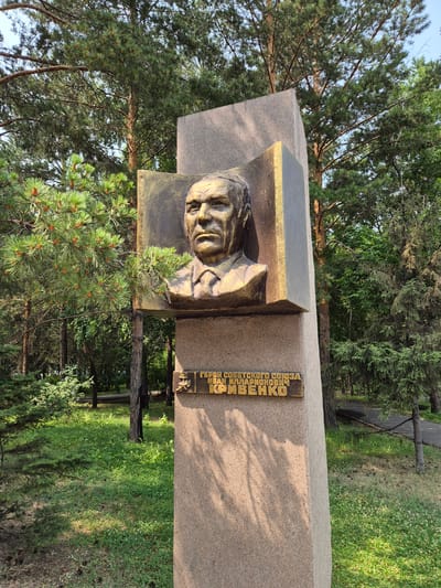 The image shows a monument in Pavlodar, Kazakhstan, dedicated to Ivan Illarionovich Krivenko, a Hero of the Soviet Union. The monument features a bronze bust of Krivenko mounted on a tall, granite pillar. Below the bust, a bronze plaque bears an inscription identifying Krivenko and his title. The monument is situated in a park-like setting with a backdrop of mature trees, including pines and firs. The ground is covered in grass and small plants, with some pinecones visible. Sunlight casts shadows across the pillar, indicating it is daytime.