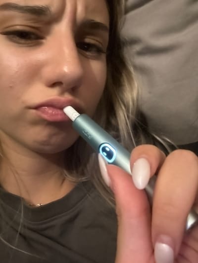 A close-up shot shows a young woman's face and hand holding a light blue electronic cigarette device. The woman has blonde hair, full lips, and is wearing a dark t-shirt. She appears to be in a casual indoor setting, possibly at home. The device is positioned near her mouth, with the tip inserted. The background is blurred, showing a neutral-colored fabric, likely a couch or blanket. No specific location in Milan, Italy is identifiable from the image.