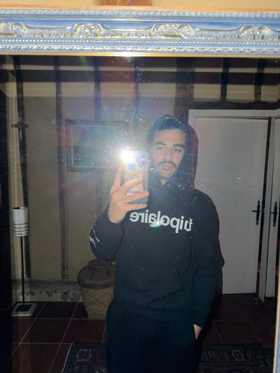 A selfie captured in a mirror dominates the image. The subject is a man wearing a black hooded sweatshirt with white reversed text, holding a smartphone. The mirror's frame is ornate, featuring a blue and gold color scheme. The background reveals a domestic interior, including a white door with black hardware, a partially visible rug with a dark pattern, brown tiled flooring, and a cylindrical object. A picture frame is also visible on the wall. The location is likely indoors, given the presence of interior design elements. The city of Mauléon-d'Armagnac, France, cannot be confirmed based on the visual information alone.