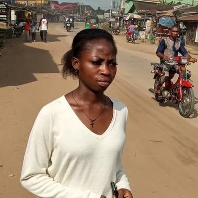 A woman in a white sweater and a cross necklace poses on a sandy street. In the background, a man drives a red motorcycle toward the viewer. Shops line the road, and other vehicles and pedestrians are visible in the distance. The image appears to be taken outdoors during the day, with a somewhat dusty atmosphere.