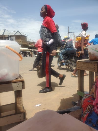 The image depicts a scene in Gwong, Nigeria, on what appears to be a sunny day, given the bright light and clear sky. The primary subjects are several people, some of whom are engaged in activities related to transportation and carrying goods.
In the foreground, a woman wearing a red headscarf and a red long-sleeved shirt is seen from the side, carrying another person, likely a child, on her back. The person being carried is wearing a patterned garment. The woman's legs are visible, wearing dark pants with red stripes.
Behind her, other individuals are present, some sitting on or near a motorcycle. One person is wearing a blue shirt and a yellow headscarf, sitting on a motorcycle. Another person, partially obscured, is wearing a blue shirt and jeans. A glimpse of a motorcycle's wheel and engine is visible, suggesting it's a mode of transport.
The setting appears to be a street or a market area. Wooden structures, possibly market stalls or seating, are present. A large translucent plastic container with an orange handle is also visible in the foreground, which might contain food or drinks. In the background, buildings with corrugated iron roofs are visible, characteristic of many structures in such locations. Utility poles and wires suggest the presence of electricity.
The overall atmosphere seems busy, with people going about their daily activities. The sunlight casts shadows, indicating it's likely daytime. There is no visible text in the image.