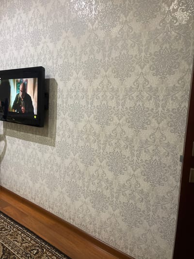 The image shows a wall with damask-patterned wallpaper and a flat-screen television mounted on it. The wallpaper is a light color with a slightly shimmering effect. The television displays a man's face. To the left of the wall, a section of a rug with intricate patterns is visible, and a wooden baseboard runs along the bottom of the wall. The lighting in the room appears to be artificial. The image is taken from a slightly low angle, looking up towards the television and the wall. There is no information in the image to indicate its location in Poltava, Ukraine.