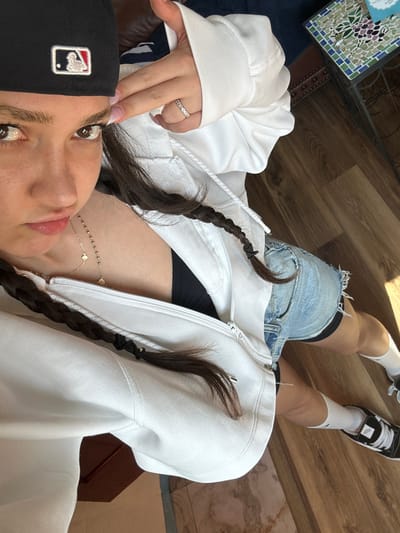 This image depicts an indoor scene in the city of Glendale, United States. A young female individual is captured from an elevated, downward-angled perspective, posing with her left hand forming a "finger gun" gesture pointed at the left side of her head. She wears a black baseball cap with a white MLB logo reversed, a white zip-up hooded sweatshirt unzipped over a black top, light-wash denim shorts with frayed hems, white socks featuring a black Nike swoosh, and black and white low-top sneakers. A thin, silver-colored ring with small stones is present on her left ring finger, and a delicate gold-colored necklace with multiple small charms is visible around her neck. Her dark brown hair is styled in two braids. The immediate environment includes light brown wood-effect flooring, with portions of a dark blue upholstered object, a brown upholstered item, and a small side table with a mosaic tile top visible in the background. Natural light illuminates the scene.