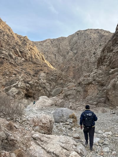 The image depicts a rocky mountain gorge in the vicinity of Quetta, Pakistan. The scene is dominated by steep, rugged, light brown and grey rock formations forming the canyon walls, which rise sharply on both sides towards a pale blue, partly cloudy sky. The ground consists of an uneven terrain of scattered rocks, boulders, and loose scree, with sparse, dry, shrub-like vegetation clinging to the slopes and ground.
In the midground, an individual is walking away from the camera, deeper into the gorge. This person wears a dark blue bomber jacket with a white "ALL STAR" logo and star on the back, dark trousers, and dark hiking footwear. A black backpack with a yellow logo is worn, and a dark baseball cap covers their head. Their posture indicates forward movement over the rocky ground.
Further into the gorge, in the background-midground, a second individual is visible. This person, smaller in scale due to distance, is partially obscured by rocks and appears to be wearing a dark green or blue jacket. They are also moving in the same direction, deeper into the canyon.
Sunlight illuminates the upper sections of the left canyon wall and the distant sky, casting shadows that suggest the sun is positioned to the upper left of the frame. The overall environment is arid and rugged, indicative of a mountainous, dry region.