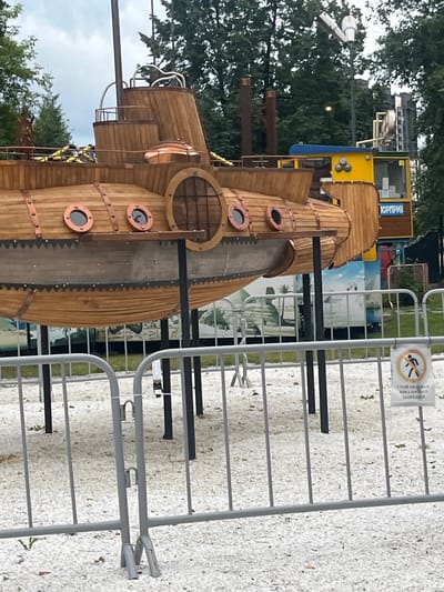 The image shows a wooden replica of a submarine, which appears to be part of an amusement park or attraction, located in Chelyabinsk, Russia. The submarine is elevated on supports and situated outdoors, surrounded by trees and a cloudy sky. In the background, a yellow structure with blue accents, possibly a kiosk or ride, is visible, along with a sign that has Cyrillic text. A metal fence with small white pebbles on the ground in front of it separates the viewer from the submarine. A sign on the fence displays a "no entry" symbol with Cyrillic text below it.