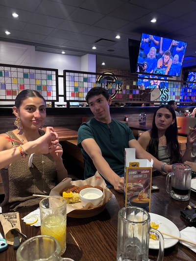 The image shows three people seated at a table inside a restaurant. The restaurant appears to be a Chili's, indicated by the "chili's" branding on the table advertising material. On the television on the wall it shows a basketball game of Golden State Warriors. Each person is looking at the camera and all 3 of them have darker hair. The table has beverages, plates, and food items that include what looks like corn and fried food with a dipping sauce. The restaurant interior includes decorative screens with colored squares, a brick wall, and flags or pennants hanging from the ceiling. From the decoration and signage, it looks like the Chili's in Puebla, Mexico.