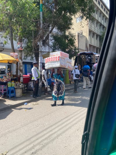 This image depicts a bustling commercial street in Accra, Ghana, likely within the central business district or near the Makola Market area, indicated by signage referencing "KAPCI GHANA LIMITED" and "PASSPORT NORTH." The scene is captured from a slightly elevated perspective, possibly from inside a vehicle, with part of a blue and black vehicle frame visible on the right.

In the foreground, a woman wearing a blue, green, and white patterned dress and black shoes walks towards the left side of the frame, expertly balancing a large, tall stack of white packaged goods (likely diapers or sanitary pads) on her head. To her left, a male security guard or traffic warden in a white uniform shirt with reflective stripes and dark trousers stands facing the street, observing the area. Behind the woman, other individuals are visible: a man in a white t-shirt and shorts, and further back, another person carrying a blue bundle on their head.

The left side of the street features an active sidewalk market. A yellow umbrella shades a stall displaying various goods, including jars, plastic containers, and packaged items on tables and colorful plastic stools. A large red cooler is positioned near this stall. Utility poles and trees line the street, with multiple signs affixed to them, one clearly displaying "KAPCI GHANA LIMITED" and another partially visible as "PASSPORT NORTH". The asphalt road surface shows some scattered litter, including plastic wrappers and a yellow object. In the background, a multi-story concrete building with numerous rectangular windows extends across the upper right, indicative of commercial or residential architecture. Further down the street on the right, additional market stalls with clothing and merchandise are visible. The scene is brightly lit by direct sunlight, casting distinct shadows.
