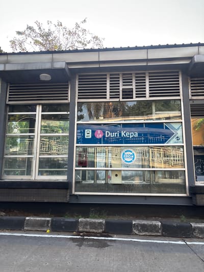 The image shows a bus stop in Duri Kepa, West Jakarta, Indonesia. The bus stop has a modern design with glass walls and a metal roof. There is a sign that says "Duri Kepa" and the number "8" and "14". There is also a "STOP NARKOBA" (Stop Drugs) sign. The bus stop appears to be empty, and there are no people visible in the image. The surroundings suggest an urban environment with trees and a street in the foreground.