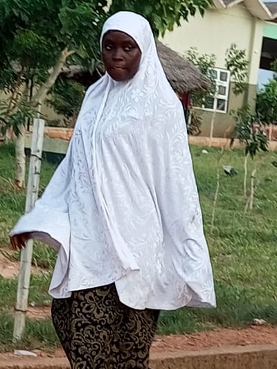 In Gombe, Nigeria, a woman stands outdoors, wearing a white hijab and a white, patterned garment that covers most of her body. Her patterned skirt is visible from the waist down. She is standing on a path with a grassy area behind her and a building with a thatched roof in the background. Two wooden posts are visible to her left. The woman is looking to her left.