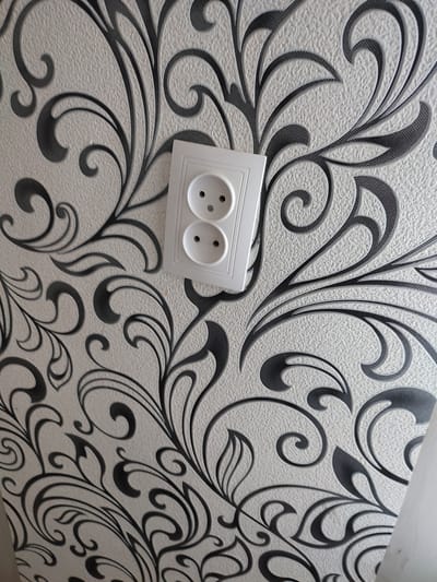 The image shows a white electrical outlet with two round sockets installed on a textured wallpaper. The wallpaper has a repeating pattern of stylized black floral motifs. The outlet is centrally located in the frame, slightly angled. The background is a light-colored, textured surface, likely a wall. The image appears to be taken indoors. There is no visible indication of the city of Narowlya, Belarus.