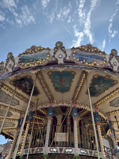 This is a low-angle, close-up shot of a carousel in Rambouillet, France, against a bright blue sky with scattered clouds and a contrail. The carousel is elaborately decorated with painted murals depicting pastoral landscapes, ships, and cherubs. Along the edge of the carousel's roof, a string of small yellow lights is illuminated. Supporting the structure are ornate columns topped with figures of mermaids with blue, scaly tails and human torsos. A sign with French text is visible on a railing within the carousel, partially obscured. The image focuses on the upper portion of the carousel's structure, revealing its intricate details and festive lighting.