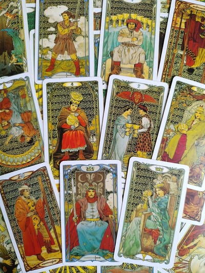 The image shows a collection of tarot cards, laid out in a fanned arrangement against a brightly lit, textured background that appears to be a table surface. The cards feature elaborate artwork in a style that suggests Art Nouveau or a similar decorative period. Each card depicts figures in historical or fantastical attire, often with symbolic imagery.
Several cards are clearly visible and identifiable. One card shows a man holding a sword, standing in a dynamic pose. Another displays a seated figure with a red turban, arms crossed, in front of a row of cups. A card with the number '4' depicts a person in regal attire holding a golden orb. The '2 of Cups' card features two figures offering each other chalices, with a lion-like creature above. Other cards show a figure seated on a throne holding a globe, another figure with a staff, and a queen on a throne. The cards are predominantly colored in earthy tones, reds, greens, and golds, with intricate black and gold decorative borders.
The image does not contain any discernible elements that would suggest a specific location within the city of Alexandrov, Russia, or any other city. The environment is confined to the presentation of the tarot cards themselves.