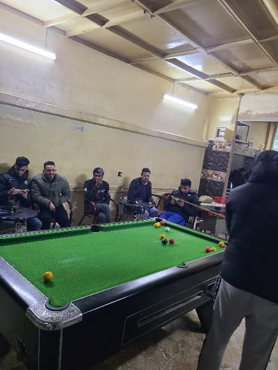 The image depicts an indoor scene in Fez, Morocco, specifically a casual establishment such as a billiard hall or café. Seven males are present. In the foreground, one male, observed from the back, wears a dark long-sleeved top and light grey sweatpants, holding a pool cue poised over a standard green-felt billiard table. The table features decorative silver-colored corners and holds multiple billiard balls, including yellow, red, and the 8-ball (black). In the mid-ground and background, six other males are seated at small tables and chairs arranged along a light-colored wall. One seated male to the left is looking at a mobile phone. Another seated male behind the billiard table, wearing a blue jacket and blue pants, also holds a pool cue and observes the game. A glass containing a green liquid is visible on one of the small tables. The room features plain light-colored walls, a grid-panel drop ceiling equipped with several fluorescent tube lights, and a patterned tiled floor. A dark-paneled partition or mirror is visible on the right side of the room.