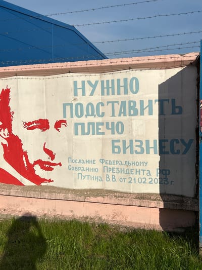 The image shows a mural painted on a concrete wall, featuring a stylized red depiction of a man's face on the left side. To the right of the face, large blue Cyrillic text reads: "НУЖНО ПОДСТАВИТЬ ПЛЕЧО БИЗНЕСУ" (It is necessary to support business). Below this main text, smaller blue text specifies: "ПОСЛАНИЕ ФЕДЕРАЛЬНОМУ СОБРАНИЮ ПРЕЗИДЕНТА РФ ПУТИНА В.В. ОТ 21.02.2023 Г." (Message to the Federal Assembly of the President of the Russian Federation Putin V.V. from 21.02.2023). The wall is topped with barbed wire, and a blue structure is visible in the background. The mural is located on the territory of the Krasnoyarsk State Agrarian University in Krasnoyarsk, Russia. The area in front of the wall is covered with green grass and shows a shadow, possibly from the photographer.
