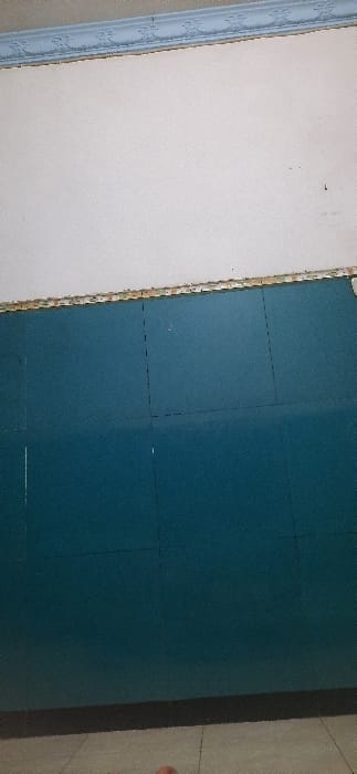 The image shows a wall in an indoor space. The wall is divided into two sections. The top section is painted white, and the bottom section is a darker blue. The wall has a small, decorative trim at the top. There is a slight discoloration in the lower portion of the blue wall, indicating a possible defect or damage. The image depicts the wall with nothing else in the frame. The location is not visible in the image, however the provided coordinates indicate it is located in Kecamatan Candi, Indonesia.
