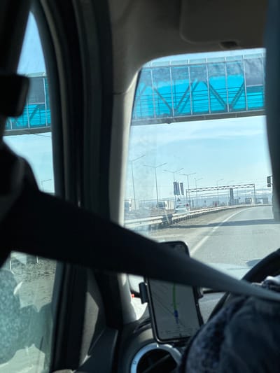 The image presents a forward-facing view from inside a vehicle on a multi-lane highway in Kulaevo, Russia. In the immediate foreground, a black seatbelt extends diagonally from the lower-left to the mid-right. A smartphone, displaying a navigation map with a green route line, is mounted on the dashboard in the lower-right, partially obscuring an air vent.

The road ahead is a paved highway with at least two visible lanes in the direction of travel, separated by a guardrail from the shoulder. Lining the road are several streetlights with distinctive angled luminaires. Overhead, a pedestrian overpass with a translucent blue panel enclosure and visible structural trusses spans the entire width of the highway. Rectangular dark-colored banners are attached to poles along the right side of the road.

In the mid-distance, a white passenger vehicle is visible on the highway, with multiple trucks and other vehicles further down the road. The surrounding environment consists of open, relatively flat terrain with light-colored ground, potentially indicating sparse vegetation or remnants of snow. The sky is clear and light blue, and a distant horizon shows subtle undulations of the land. No individuals are explicitly visible.