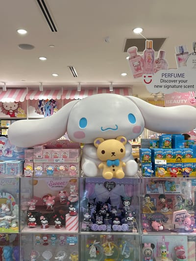 This indoor shot features a display case filled with various small toys and collectibles in Coquitlam, Canada. A large white Cinnamoroll statue with a teddy bear is prominently positioned in front of the display case. Above the statue, a sign promotes perfumes. The toy selection includes brands such as YOYO, Disney, Monsters University, Dumbo, Kuromi, Hello Kitty and We Bear Bears. The lighting is bright, typical of a retail environment.