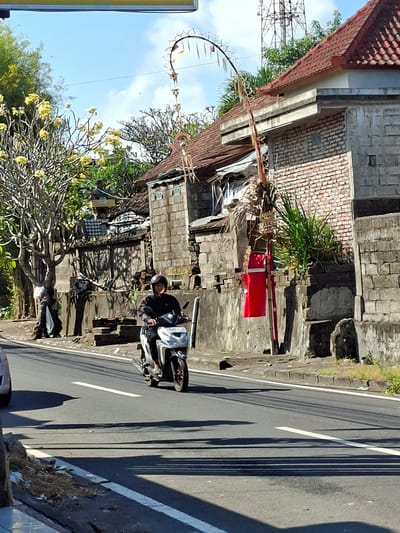 A man wearing a helmet and a dark jacket rides a white scooter down a paved road in Blahbatuh, Indonesia. The scooter has a license plate that reads "DK 4913 KN." The man is looking forward, focused on the road. The road is bordered by a concrete curb and a sidewalk. On the right side of the road, there is a brick wall, and beyond that, a building with a red-tiled roof. A tall, thin pole with decorative elements and a red banner is planted near the wall. On the left side of the road, there is a tree with yellow flowers and a stone wall, where laundry is hung. The sky is blue with some clouds.