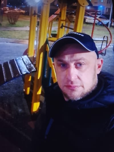 A man wearing a blue baseball cap and a black jacket is standing in front of a playground structure at night. The playground features yellow and blue metal poles and a wooden bench. In the background, there is a street with parked cars and a building. The scene appears to be in a residential area.